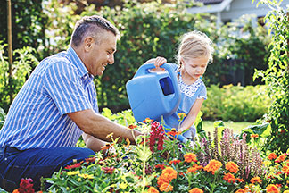 Blütenpracht: Ein Vater kniet vor einem Blumenbeet im Garten, seine kleine Tochter steht dabei und giesst mit einer blauen Giesskanne die Blumen.