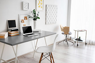 Der richtige Arbeitsplatz: Ein grauer Schreibtisch auf dem ein Laptop steht, befindet sich an einer weissen Wand an der zwei Dekobilder und eine Uhr hängen – davor ein weisser Stuhl.