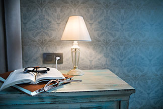 Schickes Design für Ihre Lampe: Eine Tischleuchte mit einem weissen Lampenschirm steht auf einem Tisch in Vintage-Optik, auf dem eine Mappe, ein aufgeschlagenes Buch und eine Brille liegen.