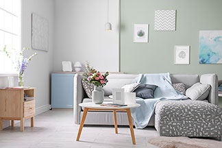 In einem Wohnzimmer steht eine grauen Eckcouch vor der ein runder Tisch mit einer weissen Tischplatte und Beinen aus hellem Holz steht.