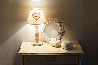 Ein rechteckiger weisser Nachttisch mit Schublade, auf dem sich zwei Keramikherzen, ein geflochtener Holzkranz sowie eine Lampe befinden, steht im Flur vor einer weissen Wand.