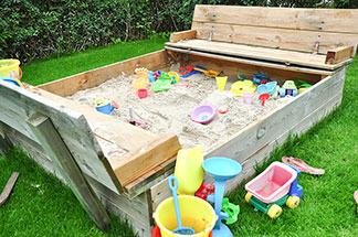 In und neben einem Sandkasten aus Holz, der auf einer Wiese steht, liegt buntes Spielzeug für draussen – darunter Sandförmchen, Schaufeln und Eimer.