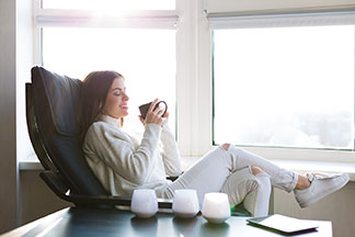 Relaxen im Sessel: Eine weiss angezogene Frau sitzt vor einer Fensterfront in einem schwarzen Relaxsessel aus Leder und hält eine Tasse Tee in der Hand.