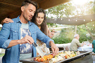 Ein junger Mann steht am Grill und wendet einen Fleischspiess mit einem Schaber, während eine junge, langhaarige Frau ihren Arm um seine Schulter legt, und ebenfalls auf den Grill schaut.