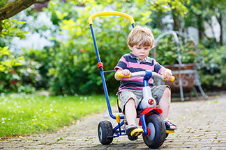 Spielzeug für draussen im richtigen Alter nutzen: Ein kleiner Junge, der kurze Hosen und ein rosa-blau-gestreiftes Shirt trägt, fährt mit seinem Dreirad über einen Plattenweg im Garten.
