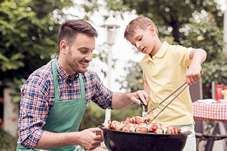 Ein junger Mann, der eine grüne Schürze trägt, kniet neben einem schwarzen, runden Grill, während sein Sohn mit einer Zange Fleischspiesse wendet.