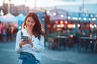 Gut erreichbar: Eine junge Frau bedient draussen ihr Smartphone, während im Hintergrund ein Strassenmarkt im Abendlicht zu sehen ist.