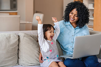 Gute Qualität lohnt sich: Eine Frau sitzt auf der Couch und hält ihren Laptop auf dem Schoss, daneben befindet sich ihre kleine Tochter, die beide Arme in die Höhe streckt und sich freut.