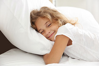Gute Qualität für guten Schlaf: ein kleines Mädchen liegt auf dem Bauch in ihrem weiss überzogenen Bett und kuschelt sich in ihr Kopfkissen.