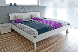 Schöne Bettwäsche als Hingucker: Ein weisses Bett, das mit gestreifter Bettwäsche in den Farben grau, weiss und lila bezogen ist.