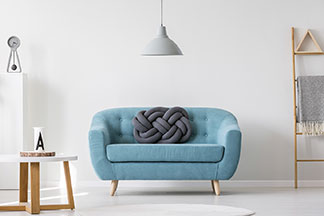 Mittig im Raum steht eine türkisfarbene Couch, über der eine eine graue Deckenlampe hängt – links im Zimmer steht ein runder Couchtisch mit weisser Tischplatte, an der Rückwand leiterförmiges Regal.