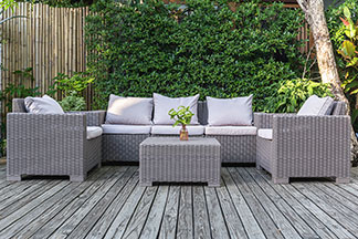Loungemöbel für den Garten: Auf einer Holzterrasse steht eine graue Couch, ein Tisch und zwei Sessel, die mit weissen Kissen dekoriert sind.