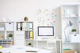 Ordnung am Schreibtisch: Ein weiss eingerichtetes Arbeitszimmer mit einem Schreibtisch, der zwischen zwei Regalen steht, in denen Ordnern und Aufbewahrungsboxen stehen.