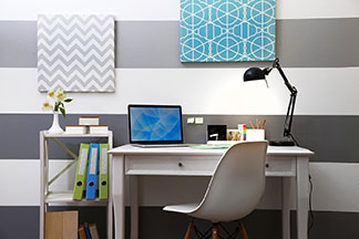 Arbeitsplatz richtig dekorieren: Ein weisser Schreibtisch mit Lampe und Monitor steht vor einer horizontal grau-weiss gestreiften Wand an der zwei bunte Dekobilder hängen.