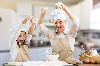 Küchentextilien zum Backen: Mutter und Tochter sind beim Backen in der Küche, tragen beide weisse Kochmützen sowie beige Schürzen und streuen mit nach oben gestreckten Händen Mehl auf ein Backblech.