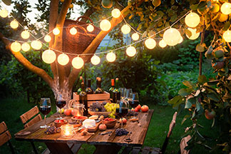 Gartenbeleuchtung als Dekoration: Ein Holzgartentisch, der unter einem Baum steht, und mit Wein, Käse und Obst gedeckt ist. Über ihm hängt eine angeschaltete Lichterkette mit runden Lampions.