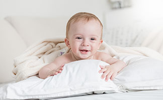 Die richtige Qualität für Ihre Kopfkissenbezüge: Ein Kleinkind liegt lachend auf dem Bauch auf einem weissen Kopfkissen im Bett.
