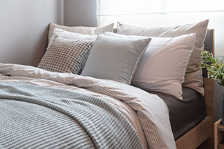Kissenbezüge aus hochwertigen Materialien: Ein Bett mit verschieden grossen Kopfkissen in hellen Erdtönen, einer gestreiften Bettdecke und einer hellblauen Tagesdecke.
