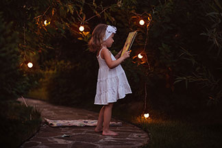 Gartenbeleuchtung in guter Qualität: Ein kleines Mädchen in einem Sommerkleid steht im Garten, der durch eine Lampionkette erhellt ist, und liest in einem Buch.