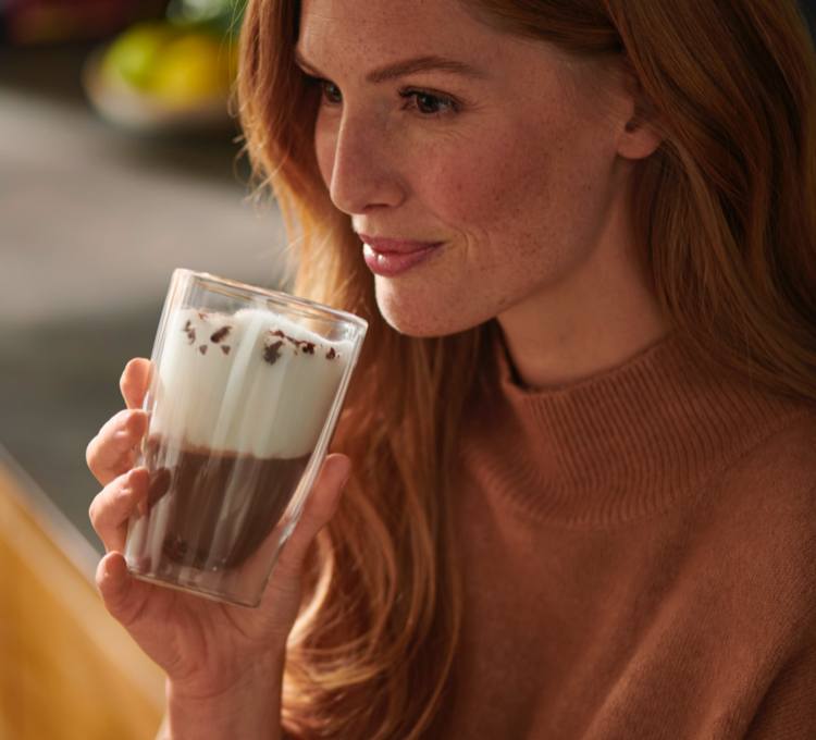 Eine lächelnde Frau trinkt einen Kaffee mit Milchschaum aus einem Latte Macchiato Glas