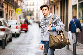 Taschen als Mode-Basic: Ein junger, modischer Mann mit Jeans und Norwegerpulli läuft mit einer grün-braunen Umhängetasche durch die Strassen einer Stadt.