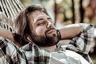 Gute Qualität bei Tchibo-Basics: Ein bärtiger Mann liegt mit weissen In-Ear-Ohrhörern und einem Holzfällerhemd in einer Hängematte und hat die Augen geschlossen.