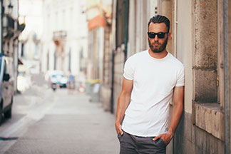 Basics für den Kleiderschrank: Ein bärtiger junger Mann mit Sonnenbrille und einem weissen Basic-Shirt steht lässig an die Mauer eines Hauses gelehnt.