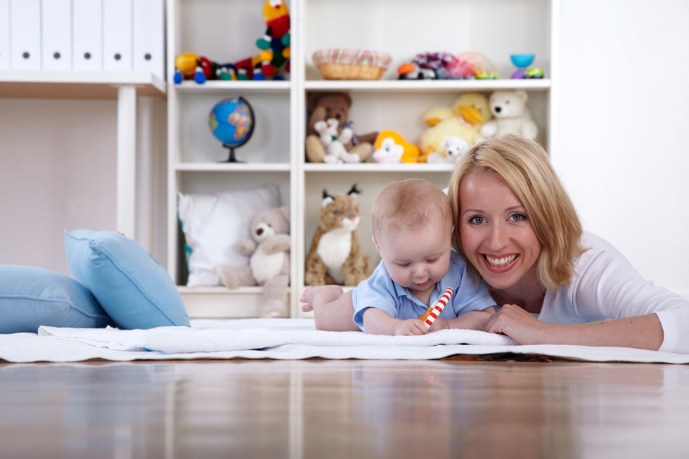 Eine blonde, lachende Frau liegt mit ihrem Baby, das mit einem rot-weiss-gestreiften Spielzeug spielt, auf einer Decke auf dem Boden im Kinderzimmer.