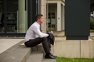 Herrenhosen fürs Büro: Ein Mann sitzt in einem weissen Hemd und einer anthrazitfarbener Stoffhose auf einer Steintreppe. Das passende Jacket hält er in der Hand.